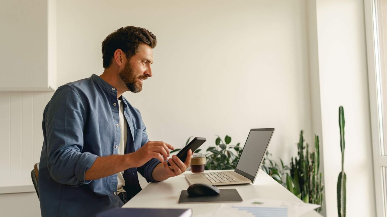 Mann nutzt ein Smartphone, während er an einem Laptop arbeitet