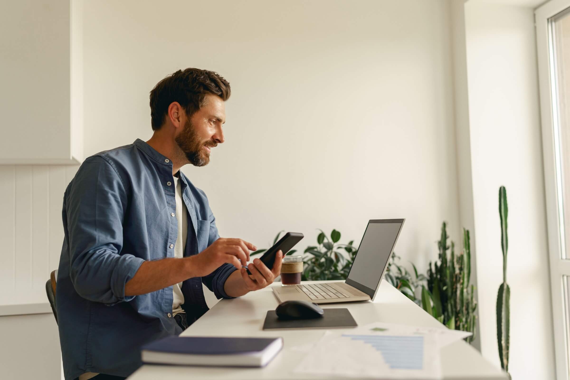 Mann nutzt ein Smartphone, während er an einem Laptop arbeitet