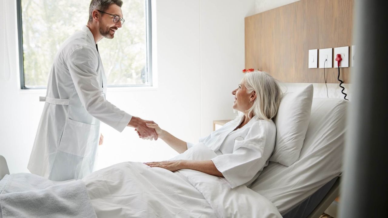 Chirurg besucht eine ältere Patientin und begrüßt sie mit einem Handschlag im Spitalzimmer