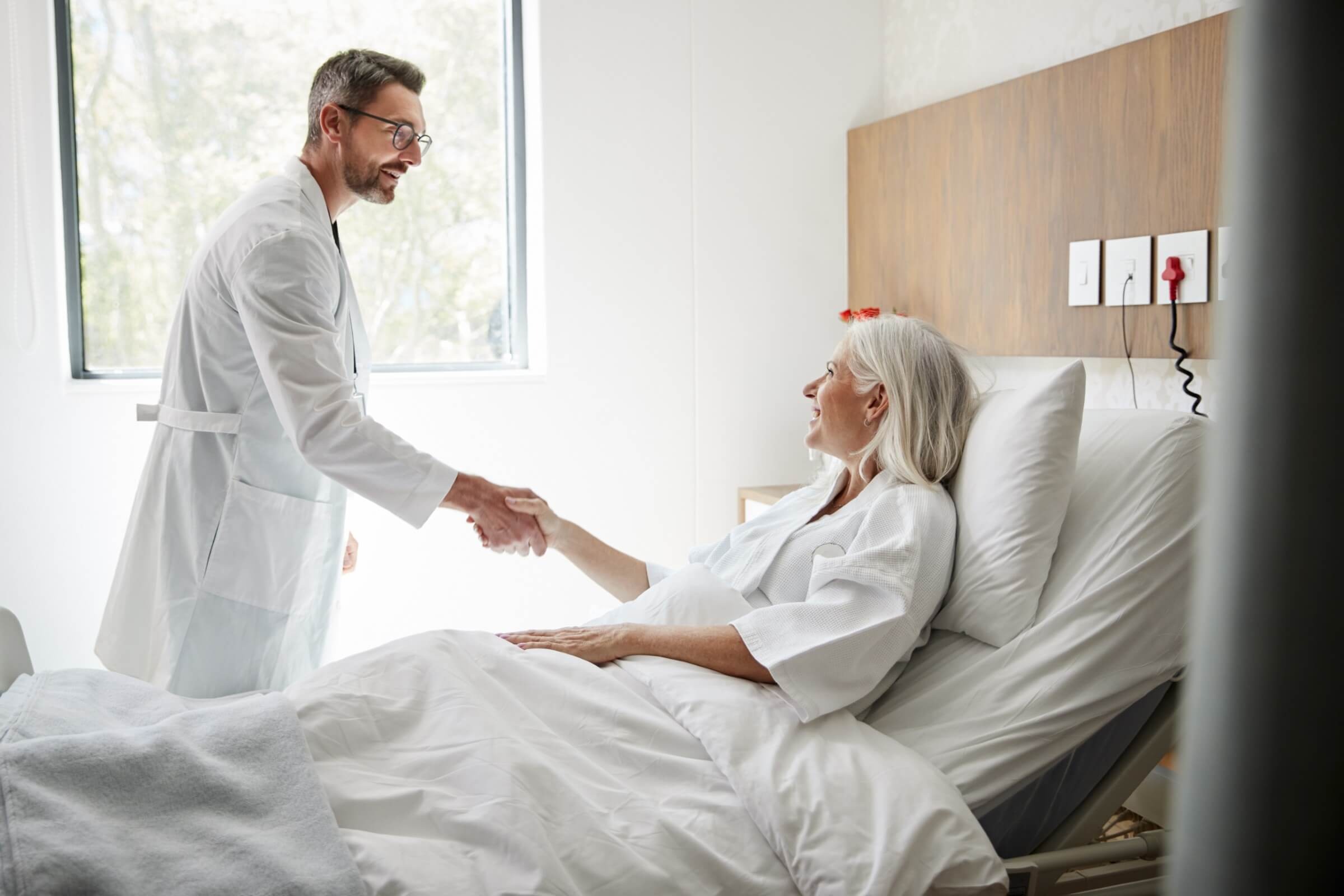 Chirurg besucht eine ältere Patientin und begrüßt sie mit einem Handschlag im Spitalzimmer